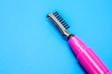 Pembe saplı ve siyah taraklı elektrikli bir kaş tarağı parlak mavi bir arkaplana yerleştirilir. Bu yakın çekim cihazın yapısını ve işlevselliğini vurgular, güzellik içeriği için idealdir.