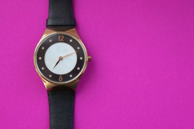 Siyah ve beyaz kadranlı zarif yuvarlak bir kol saati ve siyah deri kayışla bağlanmış altın renkli bir çanta, canlı mor bir arka plan üzerine yerleştirilir..