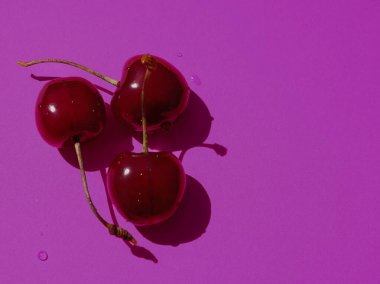 Canlı bir mor arka planda, derin bir kontrast yaratır, üç parlak, koyu kırmızı tatlı kiraz yerleştirilir, küçük su damlacıklarıyla süslenir..