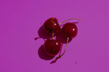 Canlı bir mor arka planda, zengin bir kontrastla, her biri küçük su damlacıklarıyla süslenmiş üç tatlı kırmızı kiraz sunulur. Bu onların doğal çekiciliğini ve çekiciliğini vurgular..