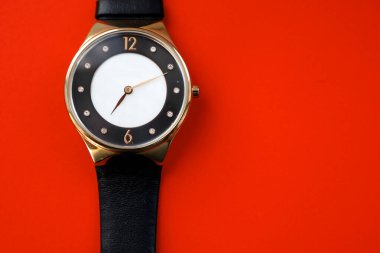 Siyah deri kayışlı yuvarlak altın bir kol saati parlak kırmızı bir arka planda yatar. Klasik dizaynı, minimalist bir kadranla zarif ve zıt bir kompozisyon yaratır..