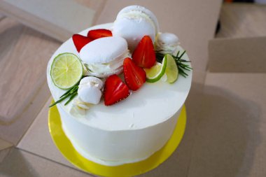 Sarı bir standın üzerinde duran beyaz bir pasta, taze çilekler, limon dilimleri, biberiye ve beyaz bezelyelerle süslenmiş. Kompozisyon hafiflik, kutlama ve tazelik hissi yaratıyor..