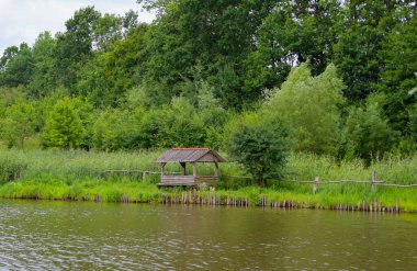 Resimli ahşap bir çardak yeşilliklerle çevrili bir su kütlesinin kıyısında duruyor. Su dalgaları doğayı yansıtır. Bir huzur ve rahatlama duygusu yaratan bir kırsal alan..
