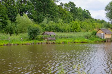 Yoğun yeşilliklerle çevrili bir su kütlesinin kıyısında küçük bir çardak ve ev. Dalgalı su yüzeyi çevredeki yaz manzarasını yansıtır..