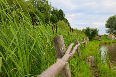 Basit bir ahşap çit sazlıklarla kaplanmış bir su kütlesi boyunca uzanır. Suyun diğer tarafında bir ahşap ev görülebilir ve kırsal bir cazibe katar..