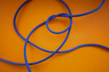 Kırmızı benekli mavi bir ip turuncu bir zemin üzerinde uzanır, bir düğüm ve çeşitli kıvrımlar oluşturur. Bu çekim halatın yapısını ve şeklini vurgulayarak, ipin dokusunu ve rengini vurgular..