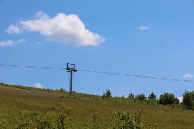 Manzaralı bir yaz manzarası, tek bir pofuduk beyaz bulutla süslenmiş koyu mavi gökyüzüne karşı kurulmuş bir kayak kaldırma kulesi olan yeşil bir yamaçtan oluşur. Parlak güneş ve açık uzay bir sükunet hissi veriyor..