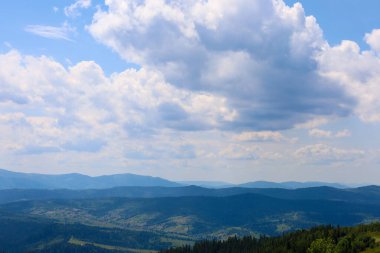 Geniş mavi gökyüzünün altında geniş, gür, gür bulutlarla dolu bir dağ sırasının manzarası. Geniş, orman kaplı tepeler puslu bir maviliğe çekiliyor, katmanlı ve geniş bir manzara yaratıyor..