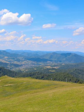 Resimli bir vadisi ve küçük bir köyü olan bir dağ silsilesinin yaz manzarası. Ön planda kuru çimenli yeşil bir tepe yer alırken, arka planda beyaz bulutlu açık mavi gökyüzünün altında sonsuz ormanlık dağlar görülüyor..