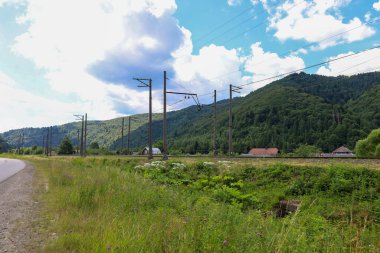 Demiryolu rayları ve elektrik hatları sık ormanlarla kaplı tepeler boyunca uzanıyor. Yakınlarda bir otomobil yolu akıyor, ve beyaz bulutlu mavi gökyüzünün arka planına karşı, küçük evler görülebilir, kırsal yaşamın bir resmini oluşturuyor.