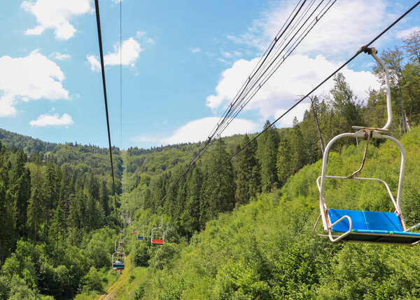 Летний горный пейзаж, где кресельный подъемник перемещается по густому лесу, открывая живописный вид на покрытые деревьями склоны. Это передает ощущение путешествий и свободы, что идеально подходит для любителей активного отдыха на свежем воздухе.