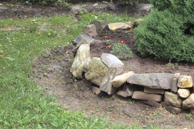 Çiçekleri yeni dikilmiş taştan bir çiçek tarlası ve dekoratif bir çalılık bahçede rahat bir köşe oluşturuyor. Manzara tasarımının bu küçük detayı doğal güzelliği ve dokuyu taşları, toprağı ve bitkileri harmanlayarak harmanlıyor..