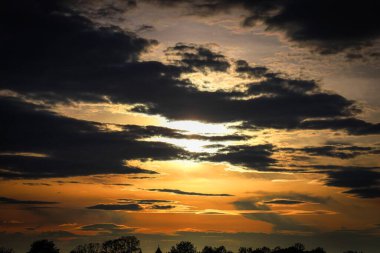 Gökyüzünde bulutların dolaştığı dramatik bir günbatımının yakın çekimi. Işıl ışıl turuncu ve altın renkler kara bulutlara karşı bir ihtişam ve sükunet atmosferi yaratır..