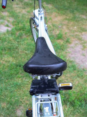 Yukarıdan aşağıya bakan bir manzara yağmurdan sonra ıslak olan eski bir bisikletin siyah deri koltuğunu gösteriyor. Eski ve biraz yıpranmış görünüyor, bu da uzun tarihini vurguluyor. Arka planda bulanık yeşil çimen ve metalik çerçeve görünür..