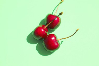 Pastel turkuaz arka planda üç olgun, koyu kırmızı kiraz yatar. Güneş ışığı dutların parlak yüzeyini vurgular ve derin gölgeler yaratır, hacim ve görsel ilgi ekler.