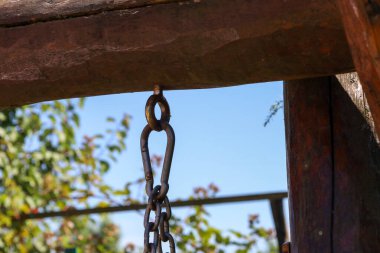 İki ahşap kirişi birbirine bağlayan devasa bir metal zincir mavi bir gökyüzü ve yeşil yaprakların arka planında asılı duruyor. Bu görüntü, kaba metal ve canlı doğayı birleştirerek bir güç ve güvenilirlik hissi veriyor..