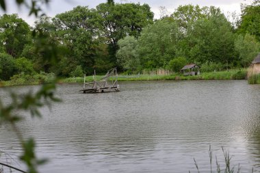 Bir göletin sakin yüzeyi, suya yansıyan yoğun yeşil bir ormanla çevrilidir. Tahta bir köprü ve kıyıda küçük bir çardak huzur ve inzivaya çekilme hissi yaratır..