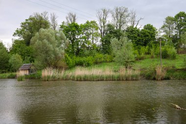 Kahverengi renkli suyu olan sakin bir gölet, kıyıdaki ağaçların yoğun yeşilliğini yansıtır. Bitkilerin arasına gizlenmiş küçük bir ahşap ev huzur ve inzivaya çekilme hissi yaratıyor..