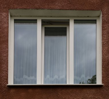 Kahverengi bir binanın ön cephesinde beyaz çerçeveli üç pencere, gri gökyüzünü yansıtıyor. Açık beyaz desenli perdeler samimi bir his katar..