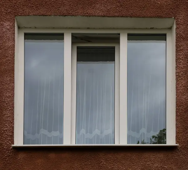 Kahverengi bir binanın ön cephesinde beyaz çerçeveli üç pencere, gri gökyüzünü yansıtıyor. Açık beyaz desenli perdeler samimi bir his katar..