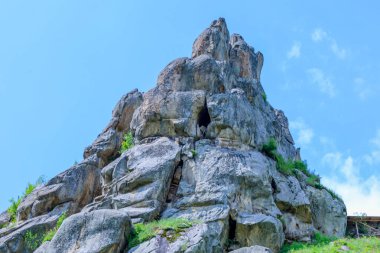 Büyük taş bloklardan oluşan kayalık bir çıkıntı açık mavi bir gökyüzünün altındaki araziye hakim. Yeşil çimen ve seyrek bitki örtüsü kırılır, taşın sertliğini yumuşatır..