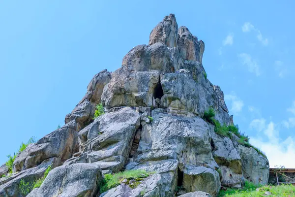 Büyük taş bloklardan oluşan kayalık bir çıkıntı açık mavi bir gökyüzünün altındaki araziye hakim. Yeşil çimen ve seyrek bitki örtüsü kırılır, taşın sertliğini yumuşatır..