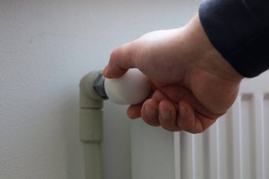 Bir insanın eli ısıtıcının beyaz termostatını oda sıcaklığını ayarlamak için ayarlar. Bu yakın çekim, ısınma mevsiminin başlangıcında sıcaklık ve enerji verimliliği üzerindeki kontrolü sembolize eder..