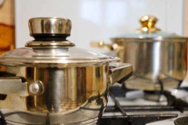 Two metal pots with shiny lids stand on the kitchen stove, the front one of which is in focus and reflects the kitchen interior. The blurred background emphasizes the homely atmosphere of cooking and the luster of the utensils.