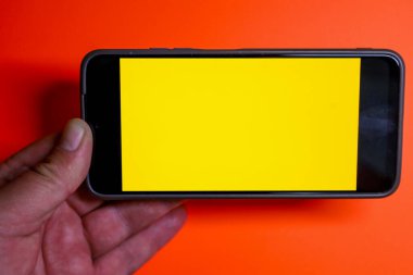Akıllı telefon bir elde yatay olarak tutulur, parlak turuncu arkaplana karşı boş bir sarı ekran görüntüler..