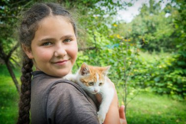 Köyde güneşli bir yaz gününde, bir kız kucağında beyaz kırmızı bir kedi yavrusu tutuyor..