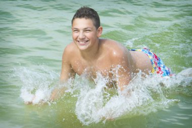 Genç bir çocuk yazın sahilde suda koşar. Her yere su sıçrıyor..