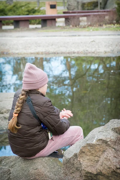 stock image In autumn, a sad girl in a hat sits on the street in a park near the water.