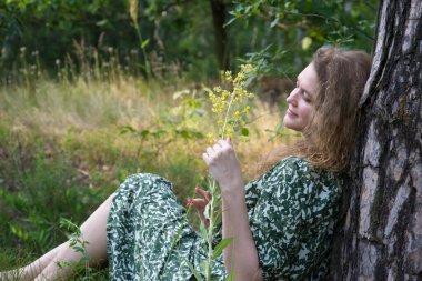 Ormanda uzun saçlı çekici bir kız. Elbise giymiş. Çimlerin altında bir ağacın altında oturuyor..