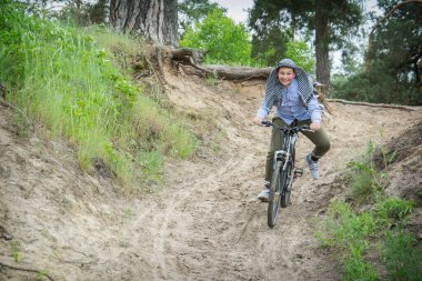 Yazın bir çam ormanında, mutlu bir çocuk dağdan aşağı bisiklet sürer. Kafasında Arapça bir başörtüsü var..