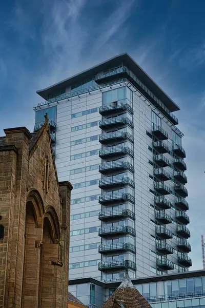 Contrast of old and new architecture with a modern glass skyscraper towering behind a traditional stone church under a clear blue sky in Leeds, UK.