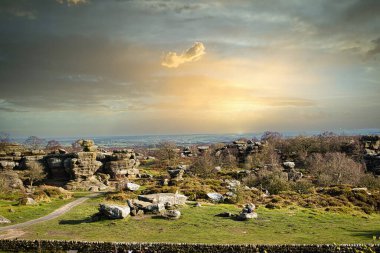 Kuzey Yorkshire 'daki Brimham Kayalıkları' nda bulutlu bir gökyüzünün altında kayalık oluşumlar ve yeşil alanlar ile gün batımında resimli kırsal alan