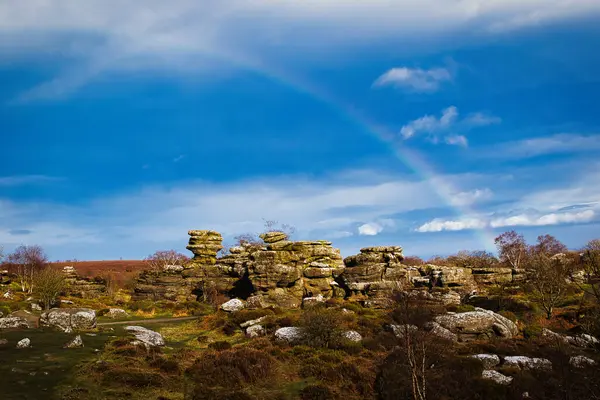 Kuzey Yorkshire 'daki Brimham Kayalıkları' nda bulutlu mavi gökyüzü altında dağınık kayalar ve yemyeşil yemyeşil kayalar üzerinde canlı bir gökkuşağı.