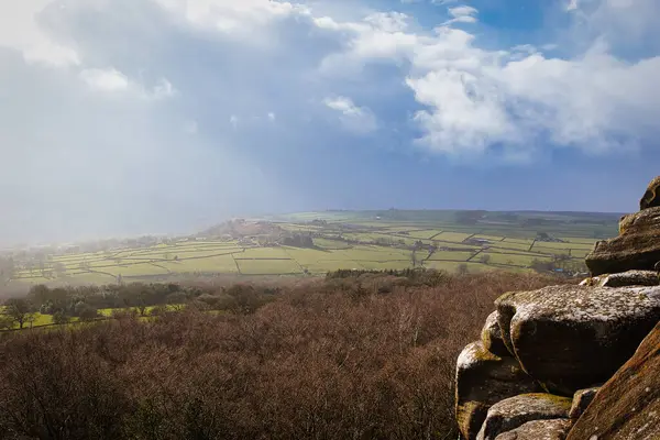 Kuzey Yorkshire 'daki Brimham Kayalıkları' nda dramatik bir gökyüzü altında kayalık önplanda güneşli bir kırsal manzaranın manzarası