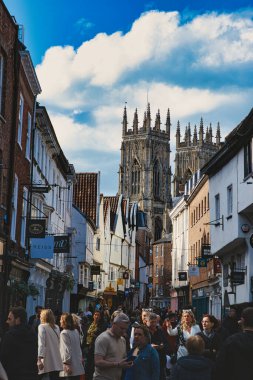 Tarihi bir şehir merkezinde yayaların bulunduğu hareketli bir sokak sahnesinde, eski binalar ve York, Kuzey Yorkshire, İngiltere 'de bulutlu bir gökyüzünün altında Gotik bir katedral yer alıyor..