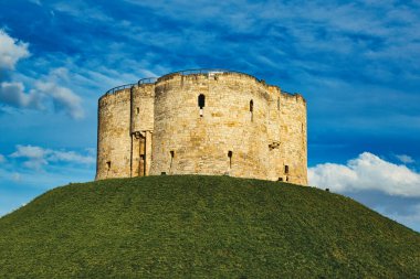 Ortaçağ taş kulesi, yemyeşil bir tepenin üstünde canlı mavi bir gökyüzüne karşı kabarık bulutlarla kaplı, York, Kuzey Yorkshire, İngiltere 'deki tarihi sağlamlığı ve antik mimariyi sembolize ediyor..