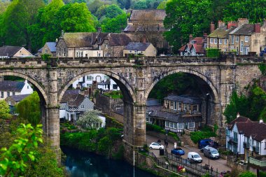Nehir üzerindeki taş bir kemer köprüsünün manzarası, yemyeşil ve büyüleyici bir köydeki antika evlerle çevrili..