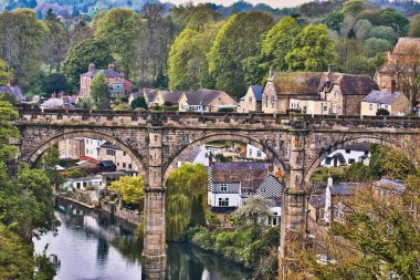 Zengin yeşillik ve geleneksel evlerle çevrili, tuhaf bir köyde nehir boyunca uzanan taş bir köprünün resimsel görüntüsü..