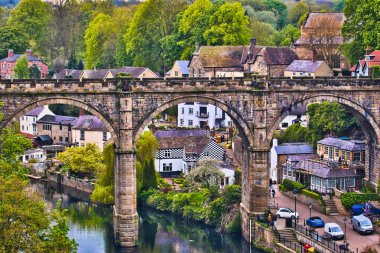 Zengin yeşillik ve tarihi binalarla çevrili, tuhaf bir köyde nehir boyunca uzanan taş bir köprünün resimsel görüntüsü..