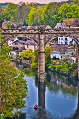 Altında yemyeşil ve antika evlerle çevrili küçük bir tekne yüzen nehrin üzerindeki eski bir taş köprünün resimsel görüntüsü..