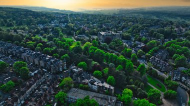 Gün batımında yemyeşil ağaçların ve evlerin olduğu bir banliyö manzarası. Manzara, uzak bir ufka sahip konut binaları ve doğal yeşillik karışımını içerir..
