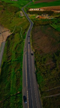 Çevresel bir yolun hava görüntüsü yeşil bir araziyi kesiyor ve üzerinde birkaç araba geçiyor. Çevrede tarlalar ve binalar var..