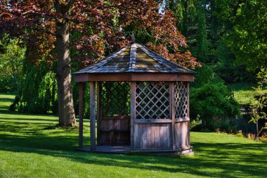 A wooden gazebo with a shingled roof in a lush green garden. The gazebo has lattice walls and is surrounded by trees and grass.