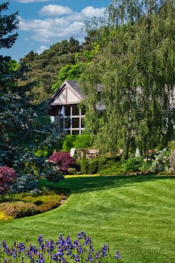 A beautiful garden with lush green grass, colorful flowers, and a variety of trees and shrubs. A wooden bench is placed near a house with a sloped roof, partially visible in the background. The sky is clear with a few clouds.