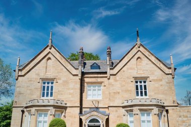 Simetrik cepheli tarihi bir taş bina, sivri kenarlar, bacalar ve kemerli pencereler. Bina ince bulutlu parlak mavi bir gökyüzüne karşı kurulmuş..