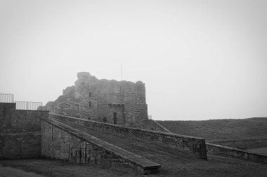 A black and white photograph of an old stone castle with a ramp leading up to it. The sky is overcast, adding a moody atmosphere to the scene.